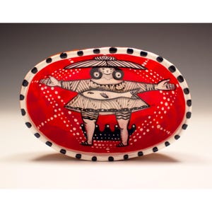 May include: Oval ceramic serving dish with a whimsical design. The dish features a red interior with a white dotted pattern and a black and white border. The central image is a figure with outstretched arms, holding a fish.