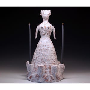 Puede incluir: Un candelero de cer&aacute;mica blanca con forma de mujer, con dos velas negras. La figura tiene una superficie texturizada y est&aacute; adornada con motivos florales y de hojas alrededor de la base. Las velas son altas y delgadas.