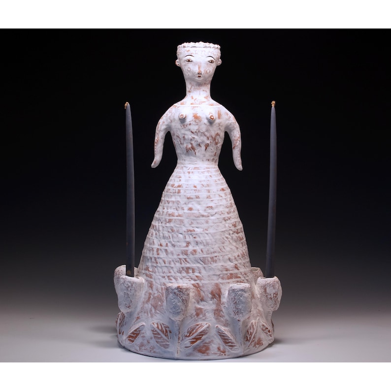 Puede incluir: Un candelero de cer&aacute;mica blanca con forma de mujer, con dos velas negras. La figura tiene una superficie texturizada y est&aacute; adornada con motivos florales y de hojas alrededor de la base. Las velas son altas y delgadas.