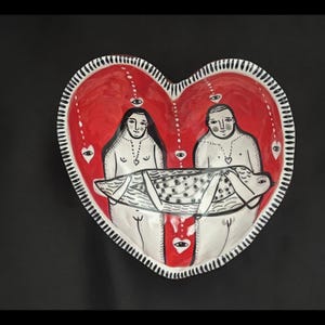 May include: A heart-shaped ceramic dish with a red interior and a white rim with black lines. The dish features two figures and a patterned object, all in black and white. The figures are facing each other, and the object is between them.