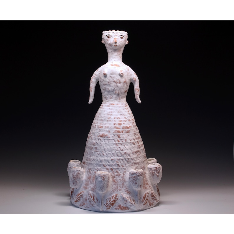 Puede incluir: Escultura de cer&aacute;mica blanca de una figura con cuello largo, brazos peque&ntilde;os y vestido texturizado. La base presenta dise&ntilde;os en forma de hojas y peque&ntilde;as estructuras en forma de jarr&oacute;n. La escultura tiene un aspecto r&uacute;stico y hecho a mano.