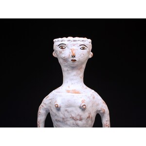 Puede incluir: Escultura de cer&aacute;mica blanca de una figura con cuello largo y rasgos faciales estilizados. La figura tiene una superficie texturizada y est&aacute; sobre un fondo negro. La escultura tiene una est&eacute;tica minimalista.