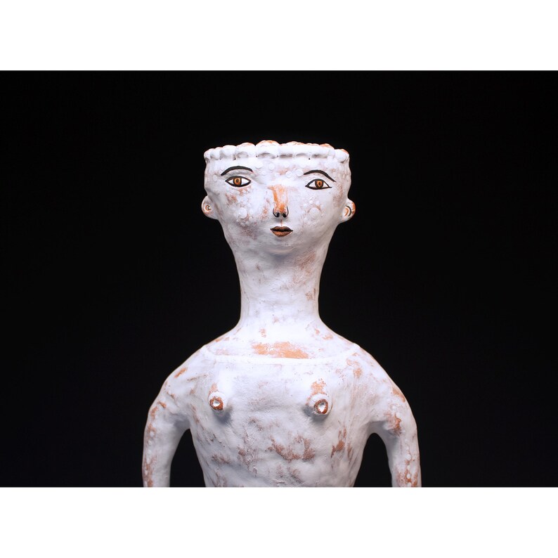 Puede incluir: Escultura de cer&aacute;mica blanca de una figura con cuello largo y rasgos faciales estilizados. La figura tiene una superficie texturizada y est&aacute; sobre un fondo negro. La escultura tiene una est&eacute;tica minimalista.