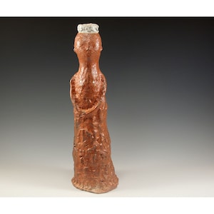 May include: A handmade, reddish-brown ceramic sculpture of a standing figure. The figure has a textured surface and a small, white, crown-like detail on its head. The sculpture is approximately 12 inches tall.