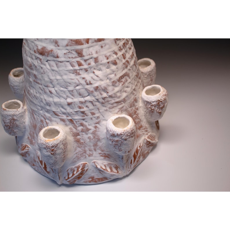 Puede incluir: Escultura de cer&aacute;mica artesanal con una superficie texturizada blanqueada. La base presenta detalles en forma de hojas y varias aberturas cil&iacute;ndricas. La escultura tiene un dise&ntilde;o &uacute;nico y org&aacute;nico, con una est&eacute;tica r&uacute;stica y terrosa.