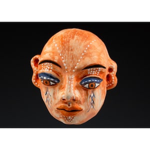 Puede incluir: Una máscara de cerámica con una base de color naranja, con rasgos faciales detallados. Los ojos están acentuados con azul y marrón, y puntos blancos y líneas azules decoran la cara. La máscara tiene una expresión serena.