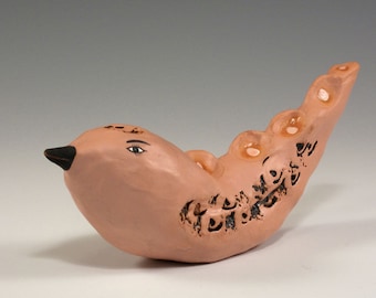 May include: A handcrafted ceramic bird figurine in a light brown hue. The bird features a black beak, painted eye details, and decorative black markings along its body. The figurine has a unique, curved shape.