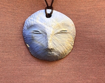 Pode incluir: Colar com pingente artesanal com um rosto estilizado com os olhos fechados. O pingente é azul claro e branco, com um cordão de couro marrom. O colar é exibido em um fundo marrom avermelhado.