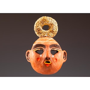 May include: A terracotta-colored ceramic mask featuring a textured, porous, beige ring-shaped ornament on top. The face has dark arched eyebrows, almond-shaped eye cutouts, small white teardrop accents, red circular cheeks, and a small open mouth.