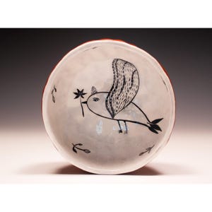 May include: A white ceramic bowl with a hand-drawn black ink illustration of a bird holding a small flower in its beak. The interior surface features a textured, uneven finish with additional small leaf motifs scattered around the bird.