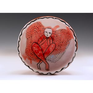 May include: A ceramic bowl with a hand-painted design. The bowl features a seated figure in red, with a red and black dotted background. The rim is outlined with a black and white wavy pattern. The bowl is a unique piece of art.