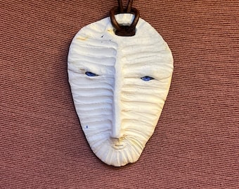 Pode incluir: Um colar com pingente branco esculpido com um rosto estilizado com olhos azuis e um cordão de couro marrom. O pingente tem uma superfície texturizada e um design minimalista, perfeito para uso diário.
