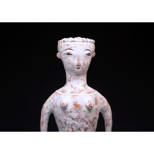 Puede incluir: Una escultura de cer&aacute;mica blanca de una figura humana estilizada con una superficie texturizada. La figura tiene un cuello largo, brazos peque&ntilde;os y un rostro detallado con ojos marrones y un tocado decorativo. La escultura est&aacute; sobre un fondo negro.