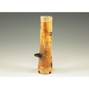 Può includere: Un vaso cilindrico alto in ceramica con una superficie strutturata, screziata marrone e beige. Il vaso presenta un occhio nero dipinto sul lato e una piccola sporgenza nera curva. La base ha dei punti neri.