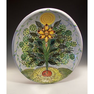 Puede incluir: Un plato de cerámica pintado a mano con un diseño central de árbol. El árbol tiene una flor amarilla con cara en la parte superior y un sol encima. El árbol tiene hojas verdes y un sistema de raíces rojas. El plato tiene un fondo azul claro y un borde decorativo.