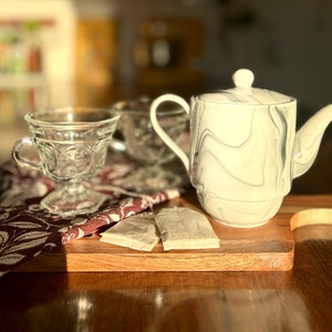 May include: A white and gray marbled teapot with a matching lid, a clear glass teacup, and two tea bags rest on a wooden cutting board. A patterned cloth and another teacup are in the background. The scene is bathed in warm sunlight.