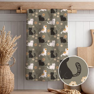 May include: A kitchen towel with a repeating pattern of black, white, and calico cats on a muted green background. The cats are interspersed with small flowers and leaves. The tail of one cat has the text "FUCK TRUMP".