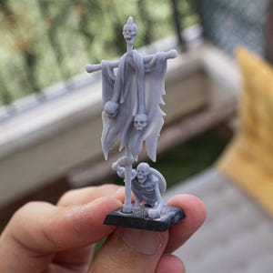 Puede incluir: Una figura en miniatura gris de un esqueleto sosteniendo un asta con una bandera con calaveras. La bandera está unida a un poste con una calavera en la parte superior. La figura está sobre una base negra.