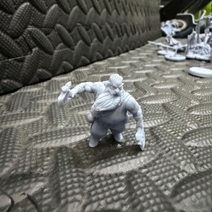 May include: A gray miniature figurine of a stout, bearded dwarf, holding a weapon in each hand. The dwarf is posed in a dynamic stance, suggesting action. The figure is set against a textured, dark gray surface, with other miniature figures in the background.
