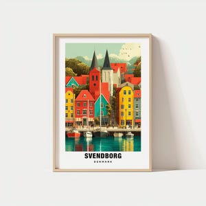 Könnte beinhalten: Ein gerahmter Druck von Svendborg, Dänemark, mit farbenfrohen Gebäuden am Wasser. Das Kunstwerk zeigt rote, gelbe und blaue Strukturen mit Türmen mit schwarzem Dach, Booten und dem Text "Svendborg Denmark."