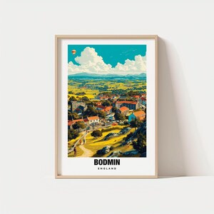 Puede incluir: Una impresión artística enmarcada de Bodmin, Inglaterra, con un paisaje colorido con un pueblo, campos y un cielo azul con nubes. Un globo aerostático flota en el cielo. La impresión está en un marco de madera clara.