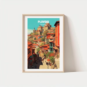 May include: A framed art print featuring a vibrant illustration of Plovdiv, Bulgaria. The artwork depicts a street scene with buildings, red rooftops, and the text "PLOVIV BULGARIA" at the top. The print is set against a light blue sky.