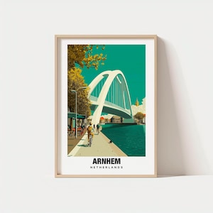 Op de afbeelding: Een ingelijste kunstafdruk met een gestileerde illustratie van Arnhem, Nederland. Het kunstwerk toont een witte boogbrug over een turquoise rivier, met fietsers en gebouwen langs de waterkant. De tekst "ARNHEM NETHERLANDS" staat onderaan.
