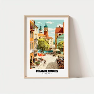 May include: A framed art print featuring a colorful street scene in Brandenburg, Germany. The artwork depicts buildings with orange, yellow, and green facades, a river, and a church with green domes. The print includes the text "BRANDENBURG GERMANY".