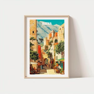 May include: A framed art print depicting a street scene in Batna, Algeria. The artwork shows colorful buildings, a mountain backdrop, and the text "BATNA ALGERIA". The print is set in a light-colored wooden frame.