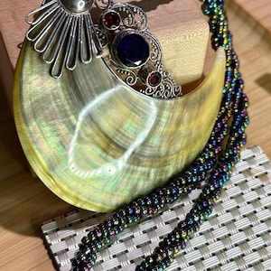 Pode incluir: Um colar com um pingente em forma de lua crescente com um design de filigrana de prata e pedras preciosas azuis e vermelhas. O pingente está preso a uma corrente de contas multicoloridas.