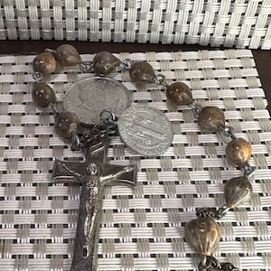 May include: A rosary with brown beads and a silver crucifix. The rosary includes two silver coins. The beads are connected by silver links. The crucifix features a detailed depiction of Jesus.