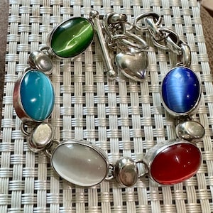 May include: A silver bracelet with a heart charm and five oval-shaped gemstones. The gemstones are green, blue, white, red, and blue.