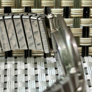 May include: Close-up of a stainless steel watch band. The band is silver and has the words "STAINLESS STEEL" engraved on it. The watch band is flexible and has a woven background.