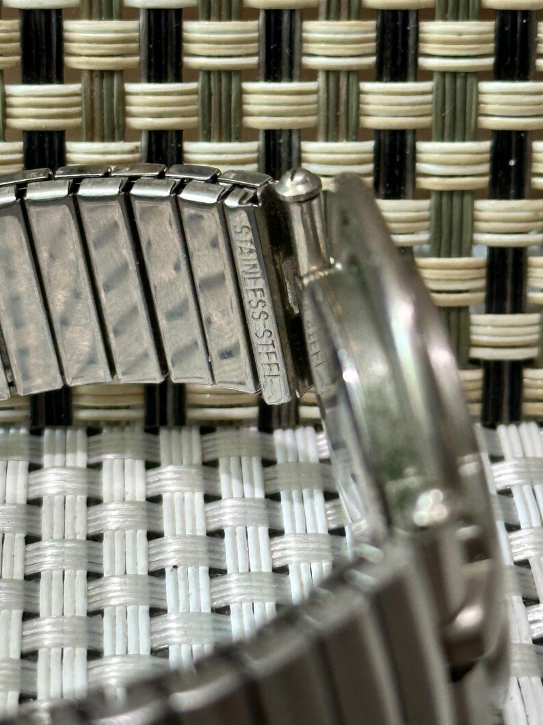 May include: Close-up of a stainless steel watch band. The band is silver and has the words "STAINLESS STEEL" engraved on it. The watch band is flexible and has a woven background.