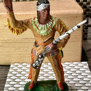 May include: A vintage toy figurine of a Native American warrior, painted in shades of brown, tan, and red. The figure is holding a rifle with a white and black pattern and is standing on a green base.