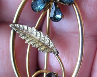 May include: A gold-toned brooch featuring a stylized blue flower with a leaf design. The flower has five petals and is set with blue rhinestones. The leaf is also set with a single blue rhinestone.