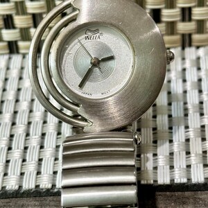 May include: A silver-toned wristwatch with a round face and a matching band. The watch face has a silver background with black hands and the brand name "WELLA". The band is made of rectangular metal links. The watch is a stylish accessory.