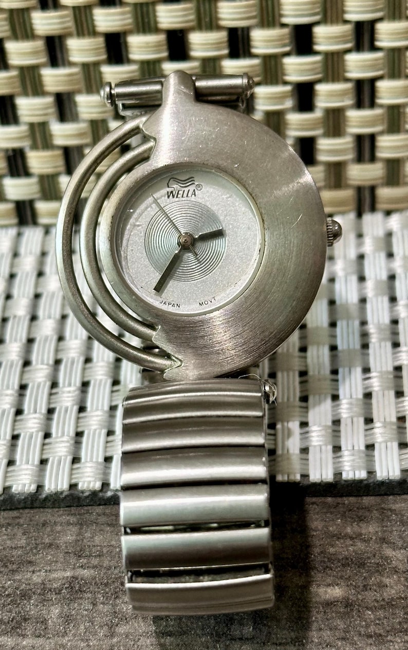 May include: A silver-toned wristwatch with a round face and a matching band. The watch face has a silver background with black hands and the brand name "WELLA". The band is made of rectangular metal links. The watch is a stylish accessory.