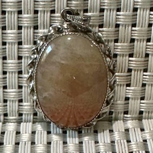 May include: A silver pendant with a light pink and white oval gemstone. The pendant has a detailed silver border.