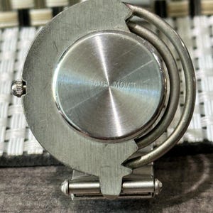 May include: Close-up of the back of a silver-toned wristwatch. The watch has a circular face with the text "PAN MOVT" and a stainless steel band. The watch is on a textured surface.