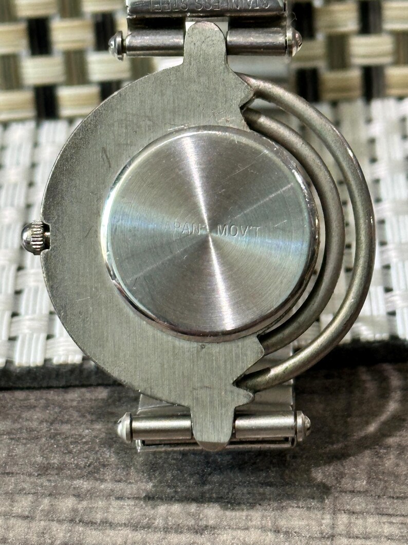 May include: Close-up of the back of a silver-toned wristwatch. The watch has a circular face with the text "PAN MOVT" and a stainless steel band. The watch is on a textured surface.