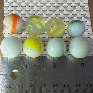 May include: Eight glass marbles in various colours, including orange, yellow, green, and blue, with swirling patterns. A metal ruler with centimetre markings is in the foreground, indicating the size of the marbles.