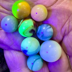 May include: A collection of eight spherical glass marbles in various sizes and colors, including bright neon green, yellow, light blue, and white, illuminated under ultraviolet light to reveal glowing surfaces and swirling patterns.