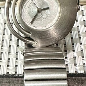 May include: A silver-toned wristwatch with a unique design. The watch face is round with a textured silver dial and black hands. The band is made of linked metal segments. The watch has the brand name "WELLA" on the face.