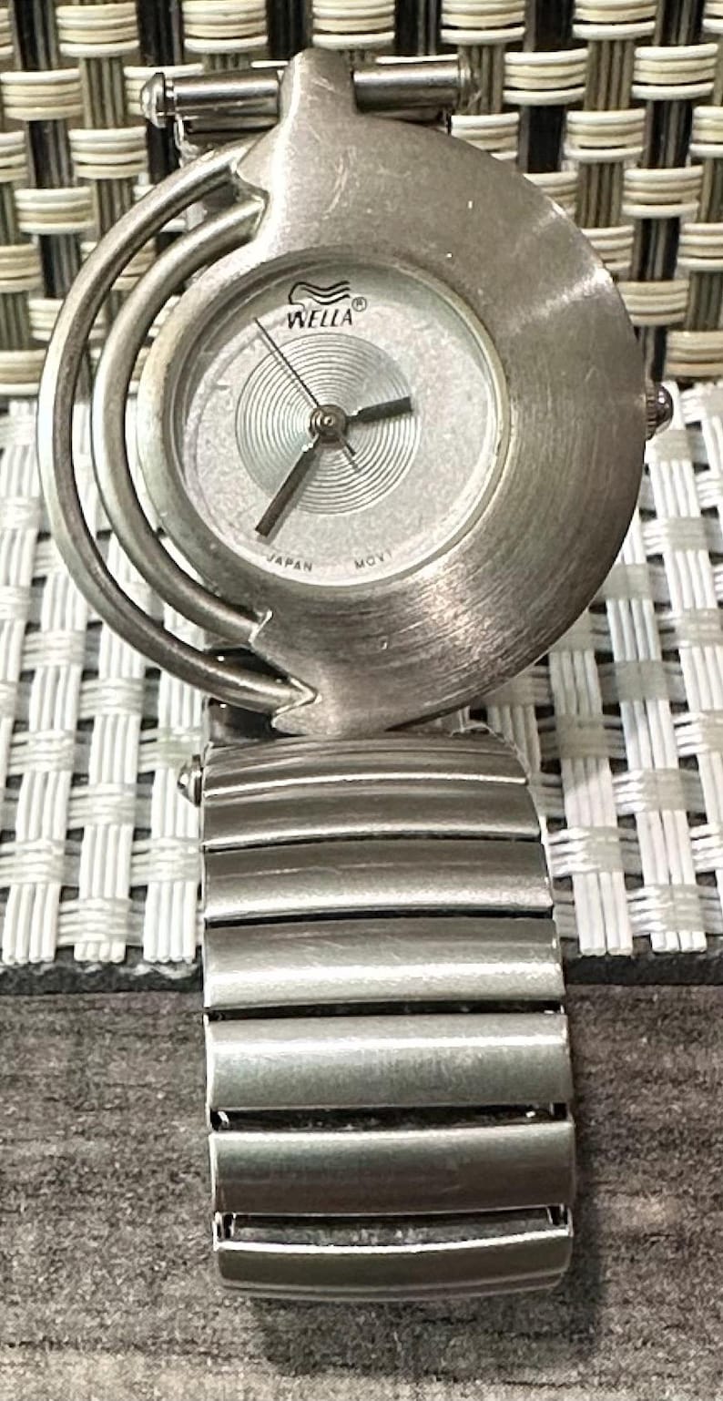 May include: A silver-toned wristwatch with a unique design. The watch face is round with a textured silver dial and black hands. The band is made of linked metal segments. The watch has the brand name "WELLA" on the face.