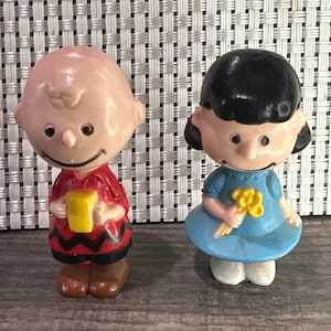 May include: Two small figurines of cartoon characters. One figure is wearing a red shirt with a black zig-zag design and holding a yellow object. The other figure is wearing a blue dress and holding a bouquet of yellow flowers.