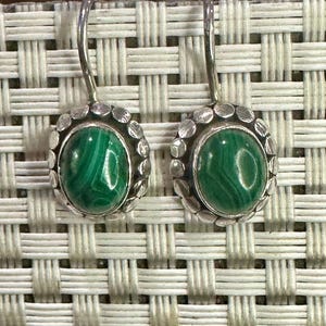 May include: A pair of silver drop earrings featuring oval, green malachite stones. The stones are set in a silver bezel surrounded by small, circular silver accents. The earrings hang from a curved silver hook.