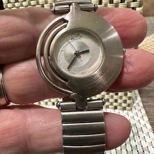 May include: A silver-toned Wella watch with a round face and a unique, curved design. The watch features a silver dial with black hands and the text "Wella" and "Japan Movt." The watch band is made of silver-toned metal links.