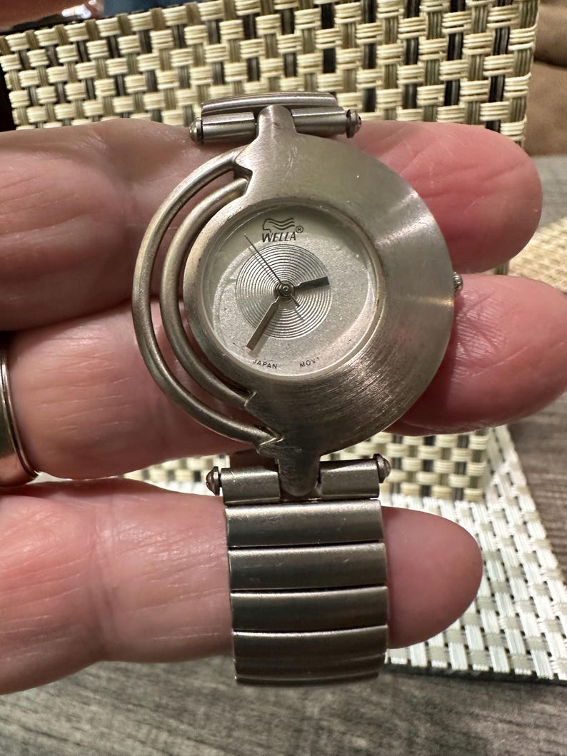 May include: A silver-toned Wella watch with a round face and a unique, curved design. The watch features a silver dial with black hands and the text "Wella" and "Japan Movt." The watch band is made of silver-toned metal links.