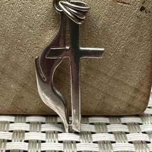May include: A silver pendant with a cross and a musical note design. The pendant has a loop at the top for a chain. The design is detailed and reflective, set against a neutral background. The pendant is likely for a necklace.